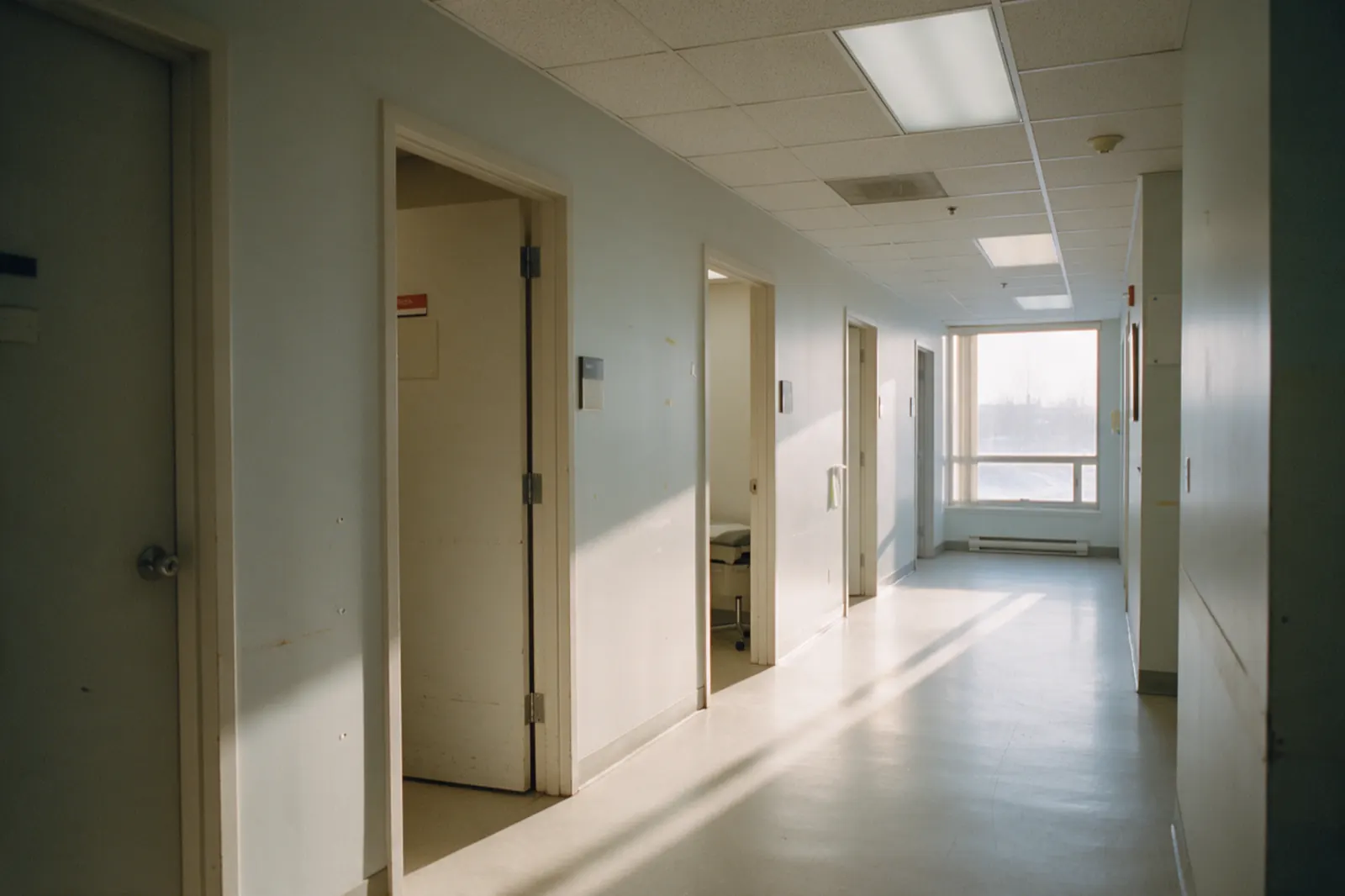 A clinic corridor stands empty, an open door showing the corner of an examination table beyond.