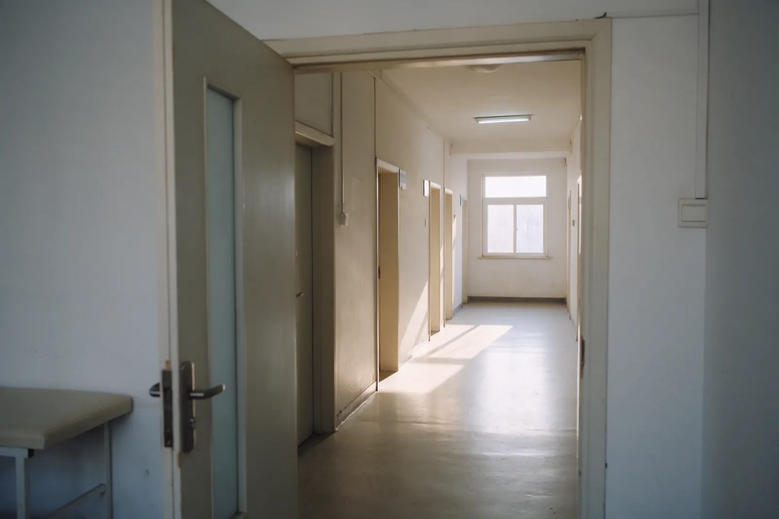 A clinic corridor stands empty, an open door showing the corner of an examination table beyond.