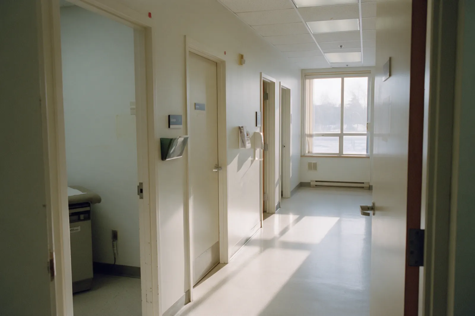 A clinic corridor stands empty, an open door showing the corner of an examination table beyond.